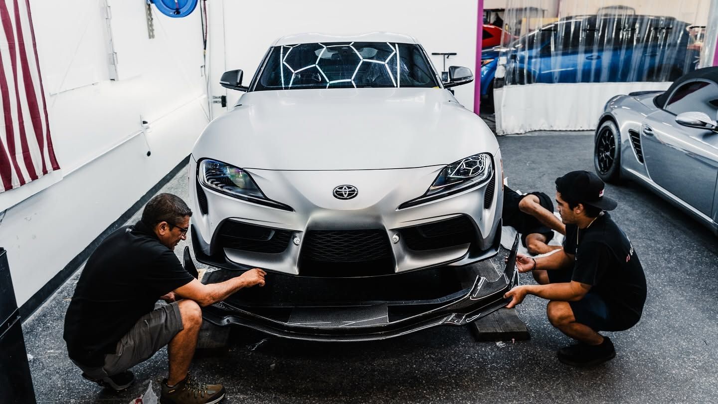 Workers detailing a white sports car in a garage, using cloths and tools on the front bumper.