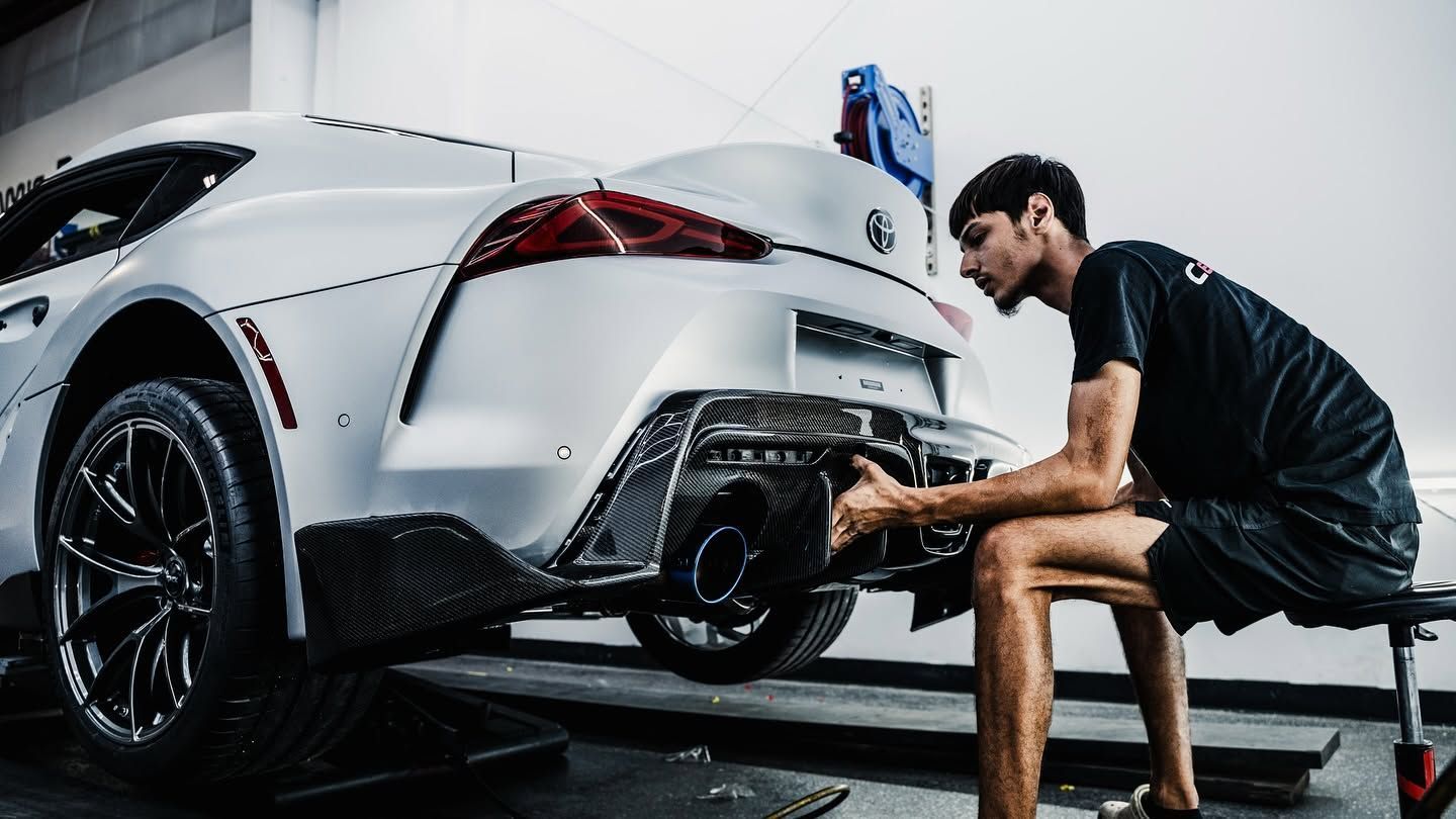 Mechanic working under a white sports car in a garage, using tools near the rear bumper.