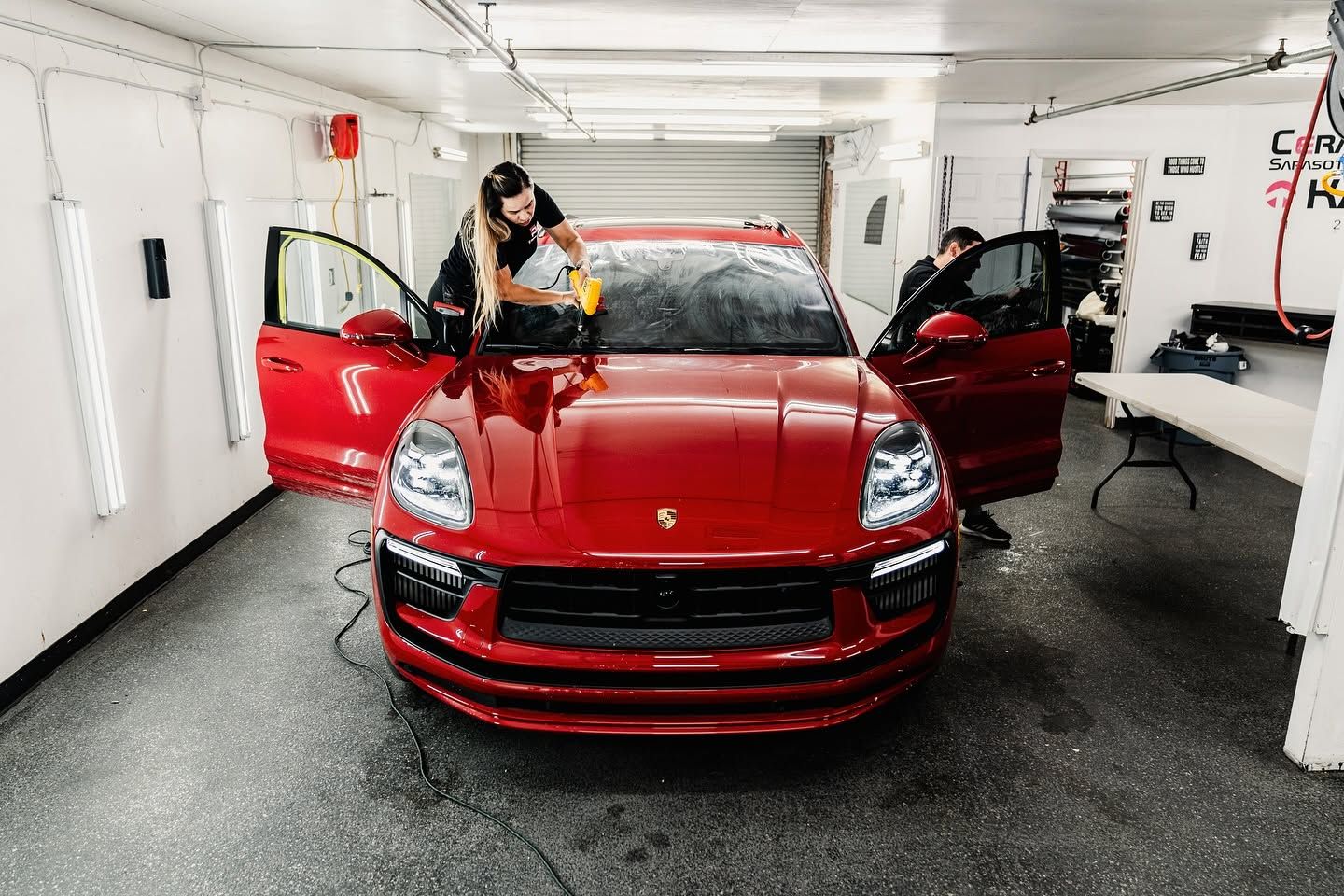 Red Porsche SUV being washed in a garage, with two people cleaning it and doors open