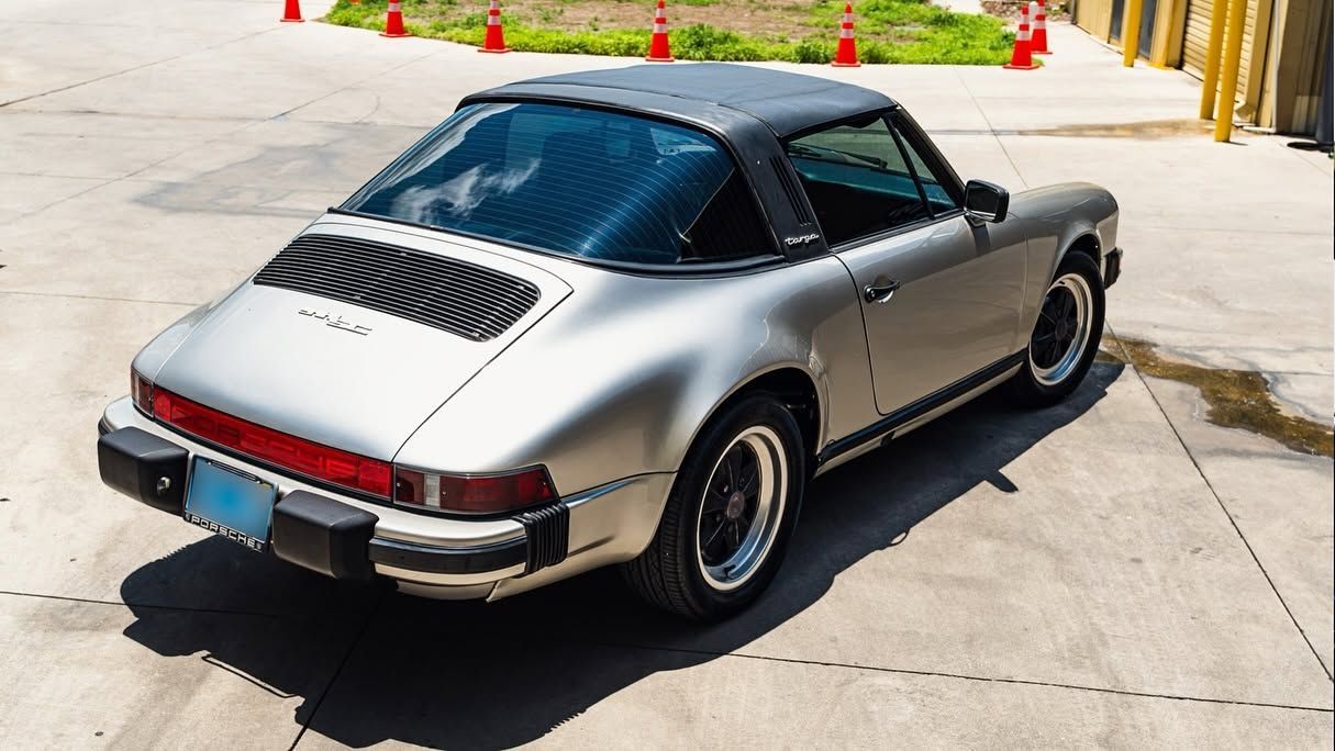 Silver vintage sports car parked on a sunlit concrete lot, rear three-quarter view with black roof