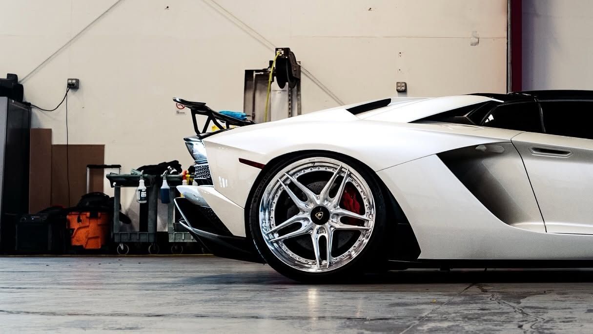 White sports car in a garage, side view with large rear wing and polished wheels.