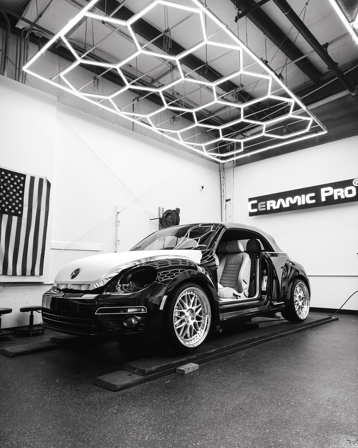 Black sports car in a garage with exposed frame, under geometric ceiling lights, beside an American flag and Ceramic Pro sign