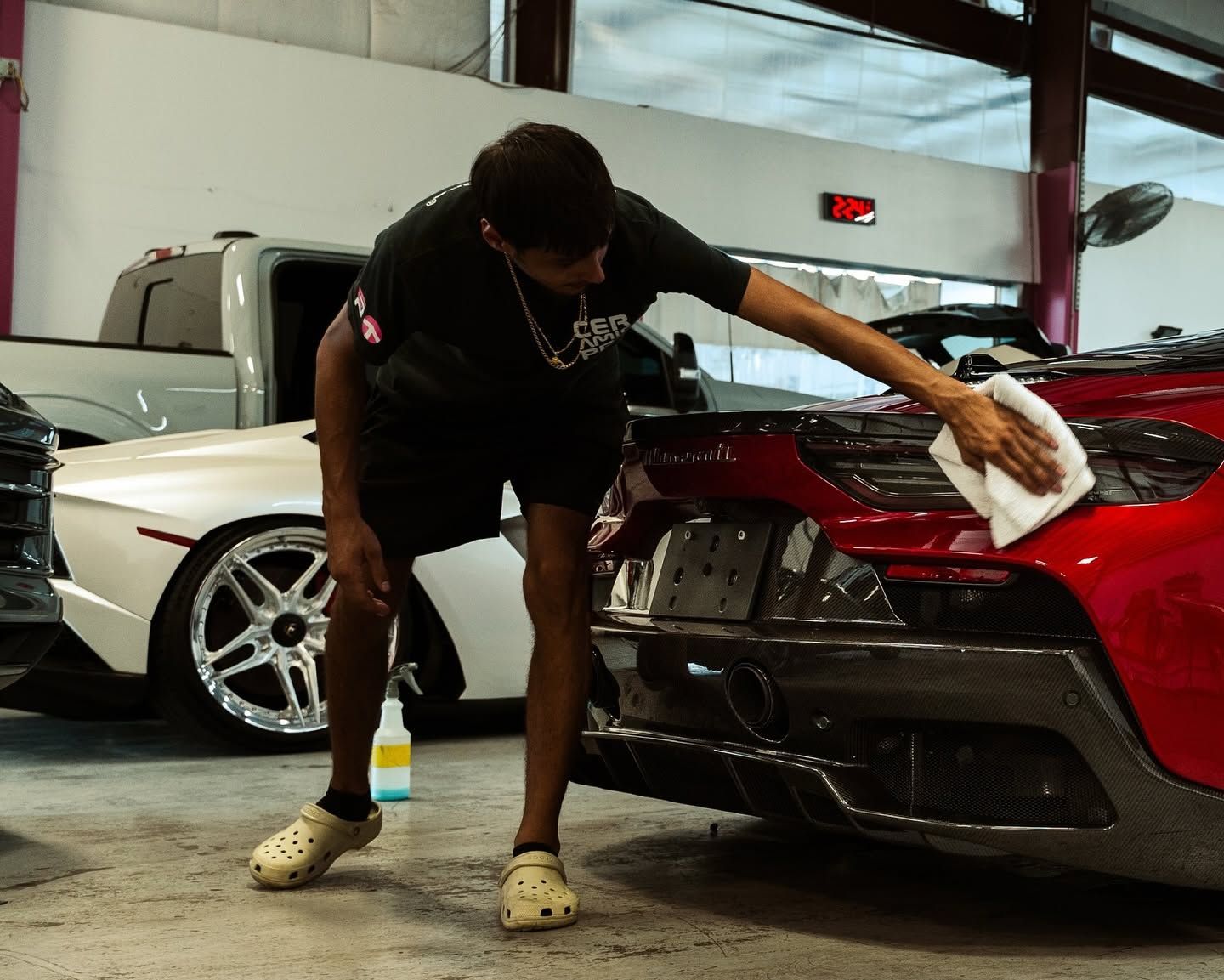 Detailing black sports car in a garage, person wiping its red rear panel with a cloth while a white car sits nearby