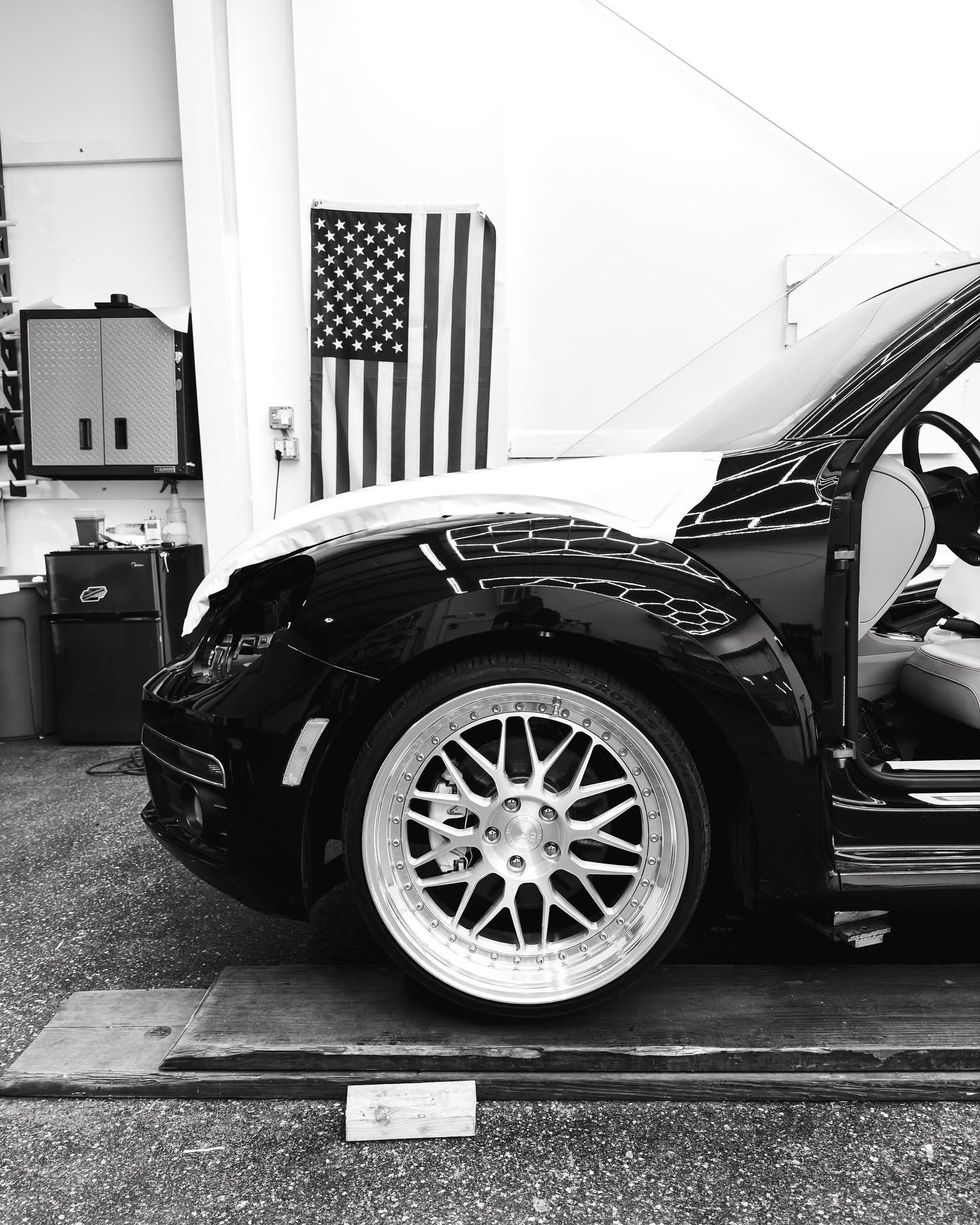 Black sports car in a garage, side view with chrome rims and an American flag on the wall.