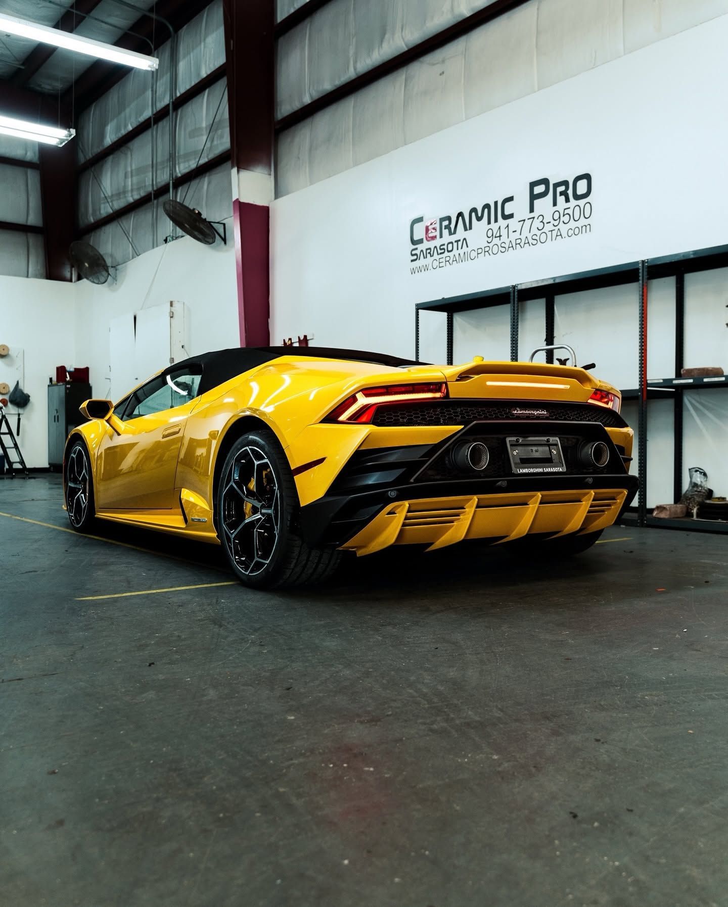 Yellow sports car in a garage, viewed from the rear with black accents and large spoiler.