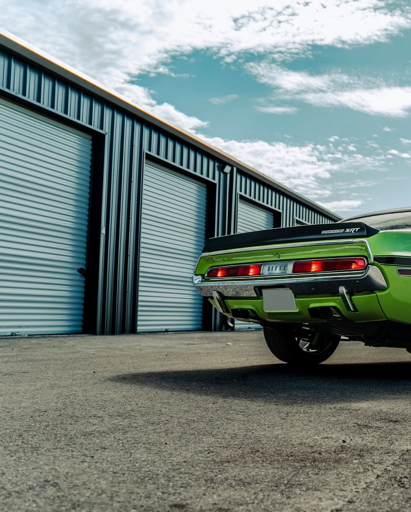 Green classic muscle car parked beside blue industrial garage doors under a partly cloudy sky