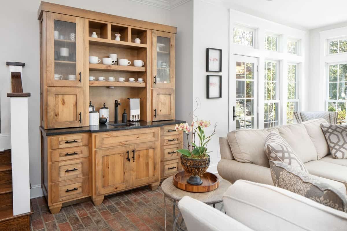 Wooden hutch with dishes in a bright living room beside a beige sofa and sunlit windows