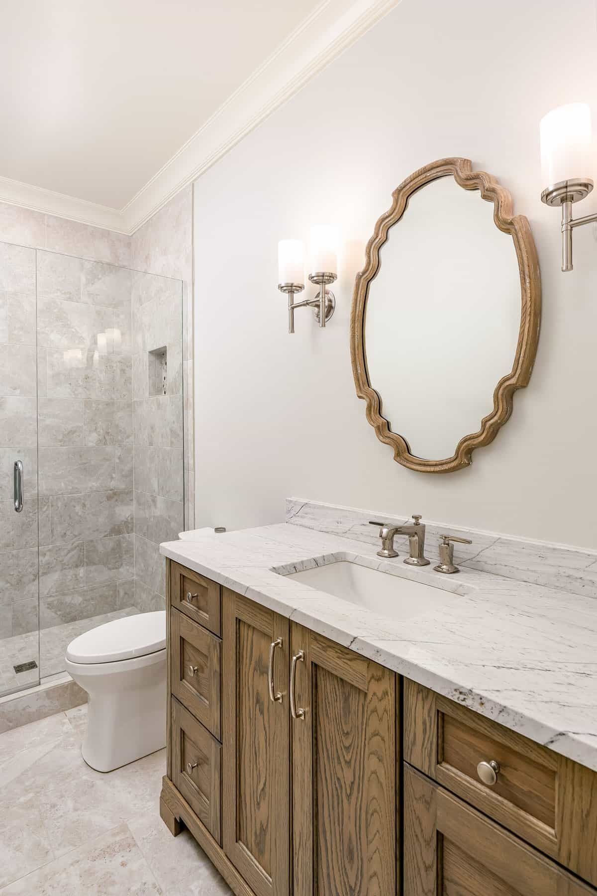 Elegant bathroom with marble vanity, oval gold mirror, wall sconces, toilet, and glass shower.