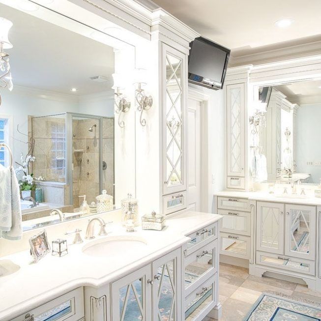 Elegant white bathroom with dual vanities, large mirrors, a glass shower, and a wall-mounted TV