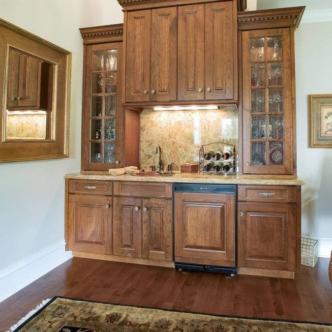 Built-in wooden wet bar with cabinets, glass-front shelves, granite counter, and tile backsplash in a living room.