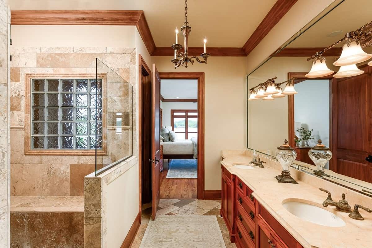 Spacious beige bathroom with double vanity, large mirror, glass shower, and doorway to a bedroom.
