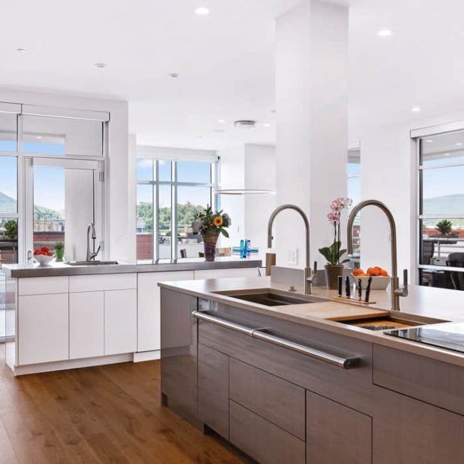 Bright modern kitchen with white cabinets, wood floors, large windows, and a central island with sink and faucet