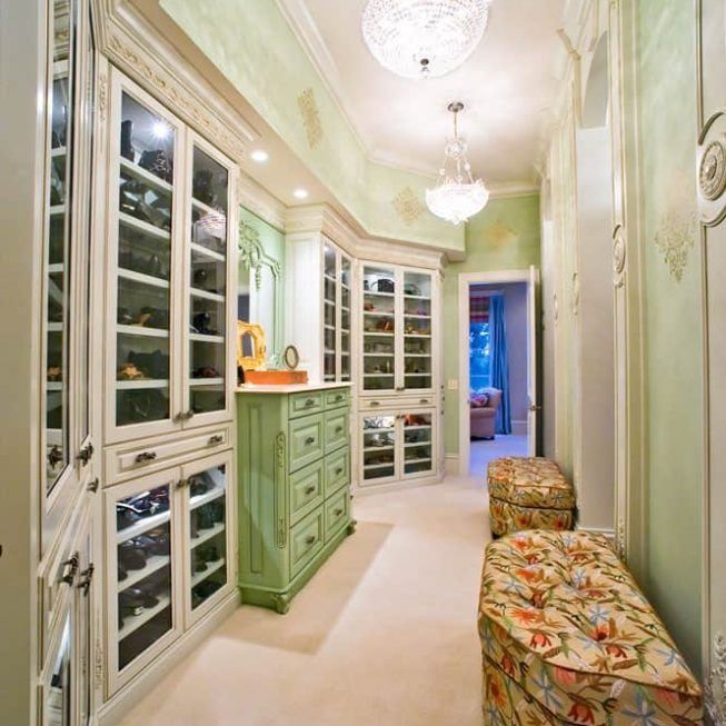 Elegant narrow walk-in closet with mint-green cabinets, glass-front shelves, chandeliers, and floral bench seating