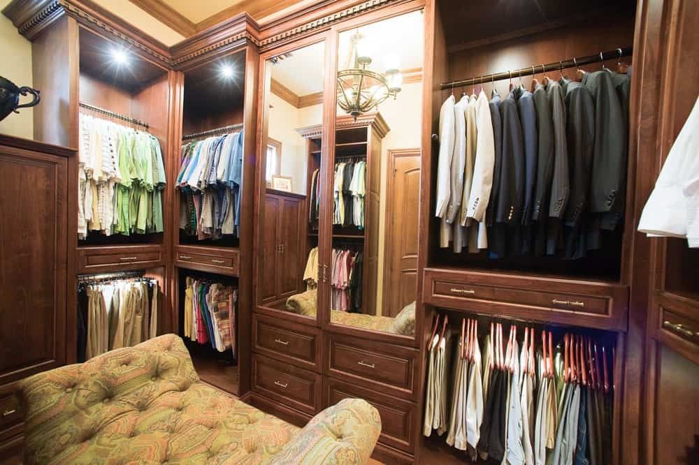Luxury walk-in closet with wood cabinetry, hanging clothes, mirrored doors, and a tufted bench.