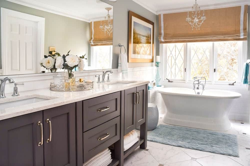 Bright bathroom with dark vanity, white marble counters, freestanding tub, and large windows with woven blinds