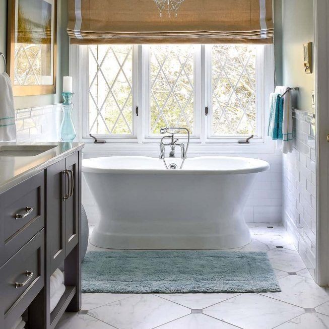 Bright bathroom with a white freestanding tub, large window, gray vanity, and blue rug