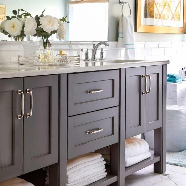 Gray bathroom vanity with double sinks, chrome faucets, white countertop, and folded towels below