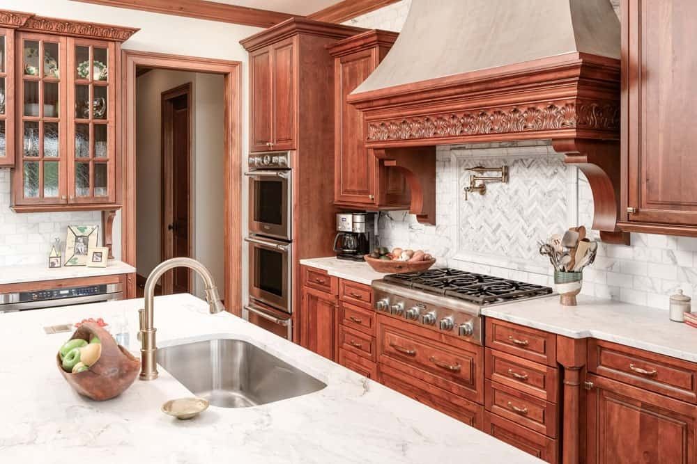 Wooden kitchen with ornate cabinets, marble island, stainless sink, and large range hood.