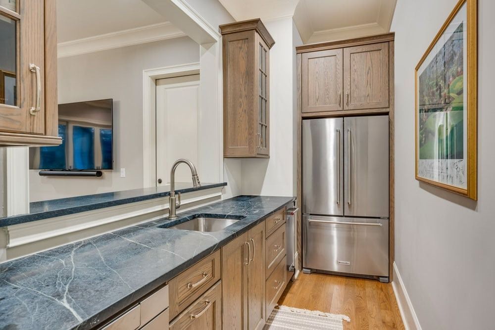 Modern kitchen with gray countertops, wood cabinets, stainless steel fridge, and a sink by a pass-through window