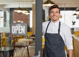 Portrait of smiling owner standing at his restaurant gate with open signboard