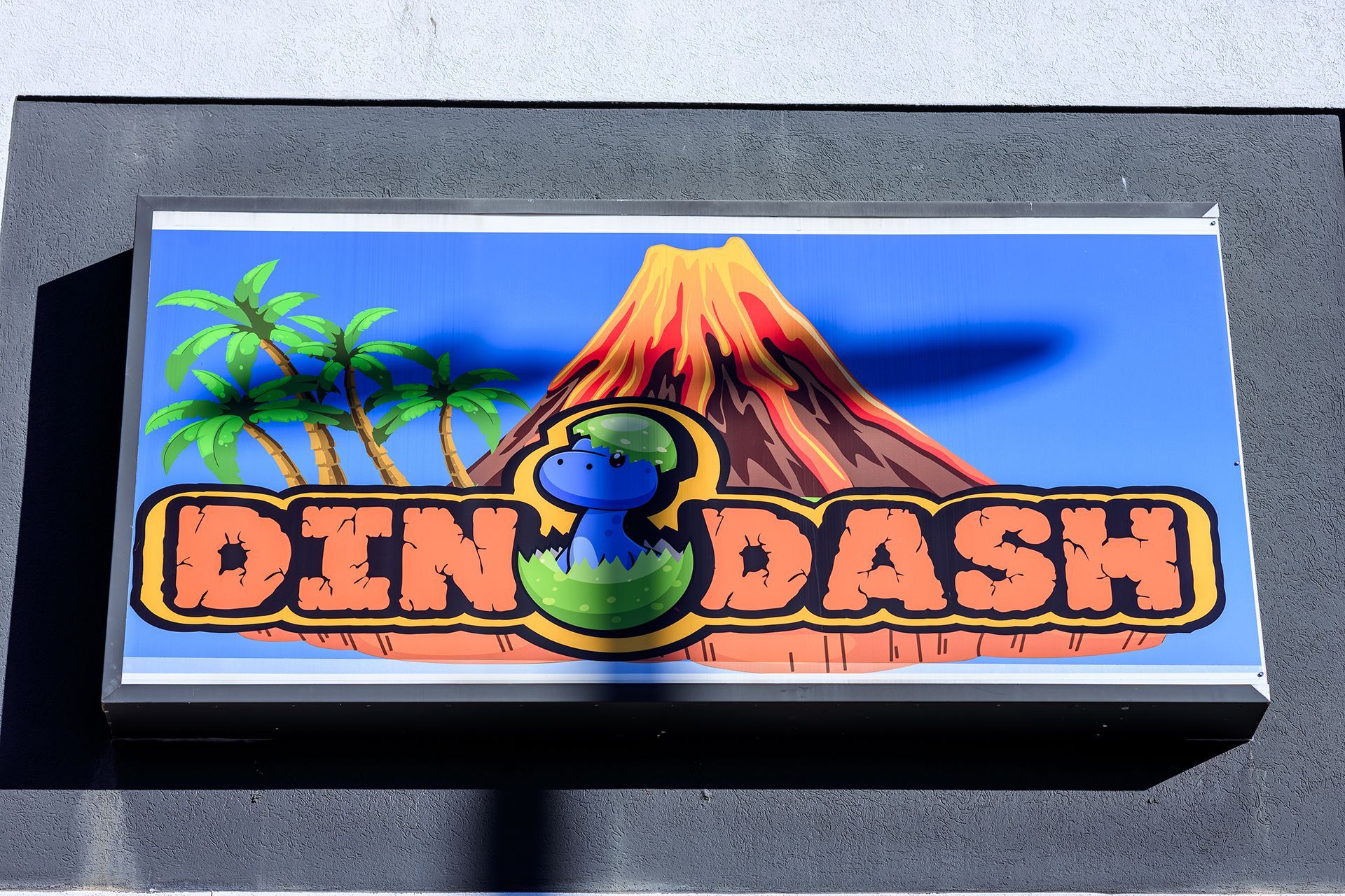 Sign for DinoDash with a blue dinosaur hatching from an egg, volcano, and palm trees.
