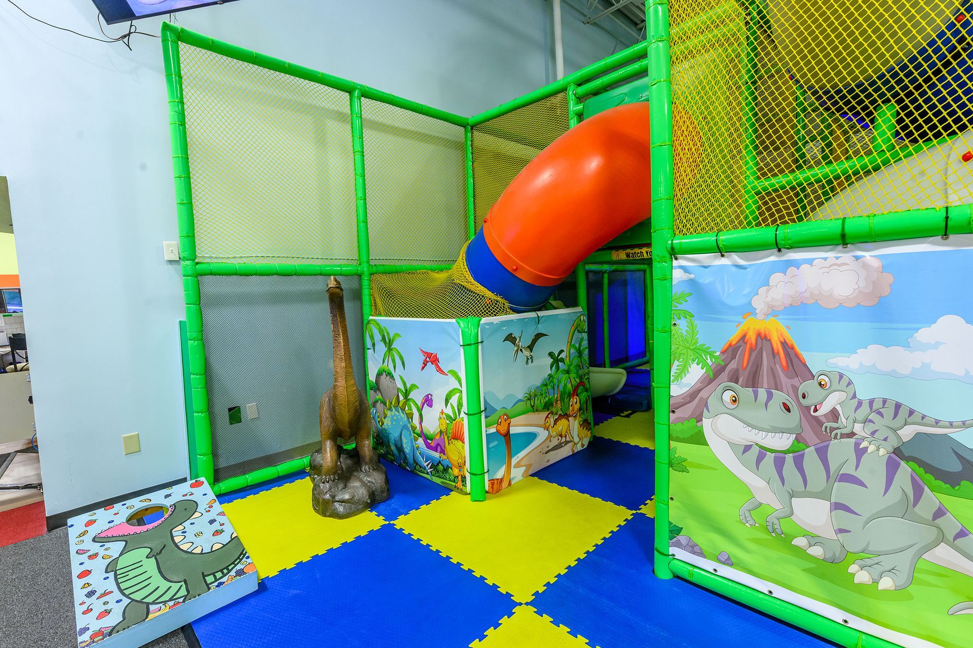 Dinosaur-themed indoor playground with slide, netting, colorful mats, and dinosaur illustrations on walls.
