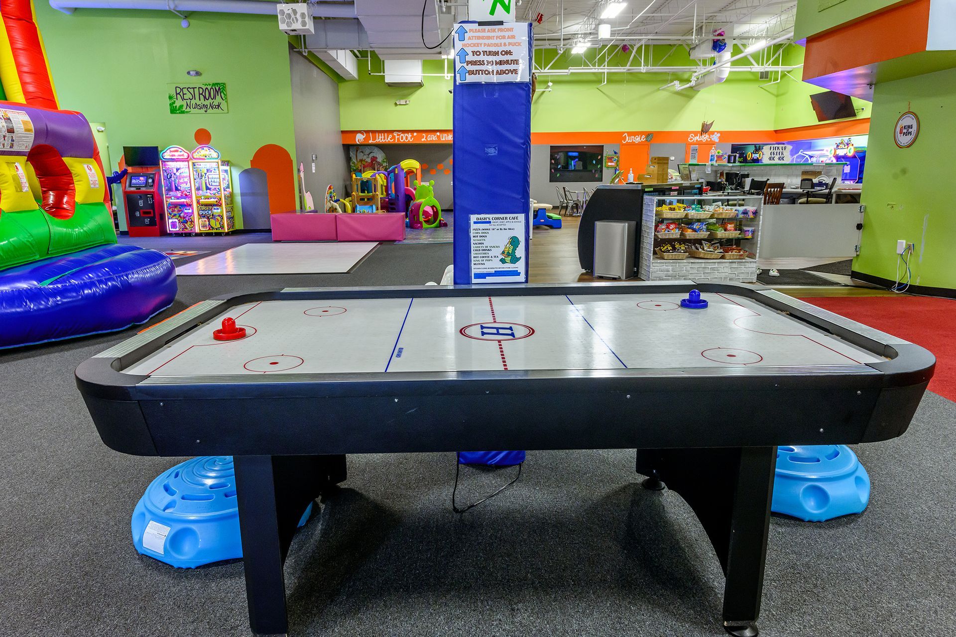 An air hockey table in a brightly colored indoor play area, with arcade games and inflatable slides in the background.