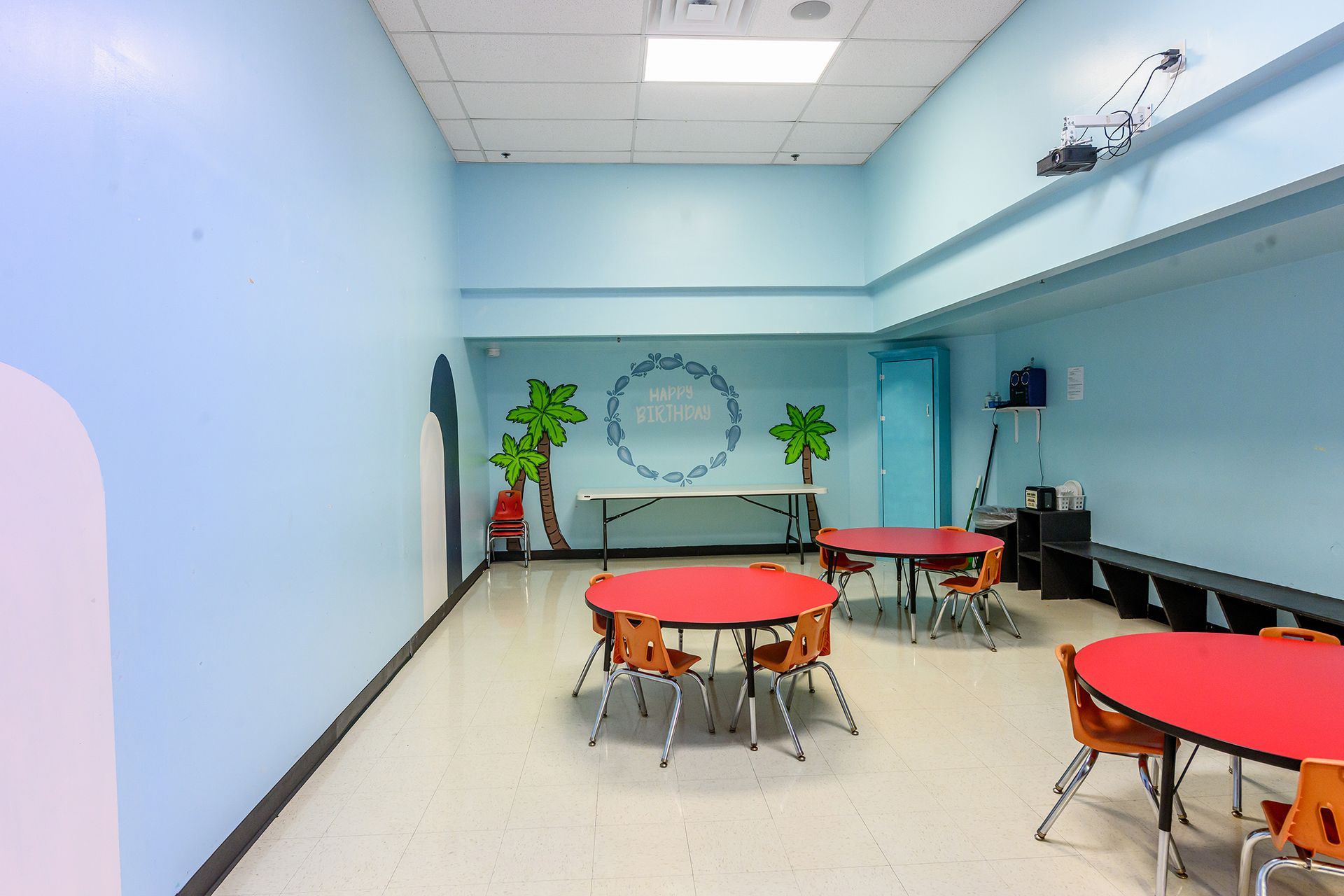 A classroom with blue walls and red tables. Palm tree mural, chairs, and a table are also visible.