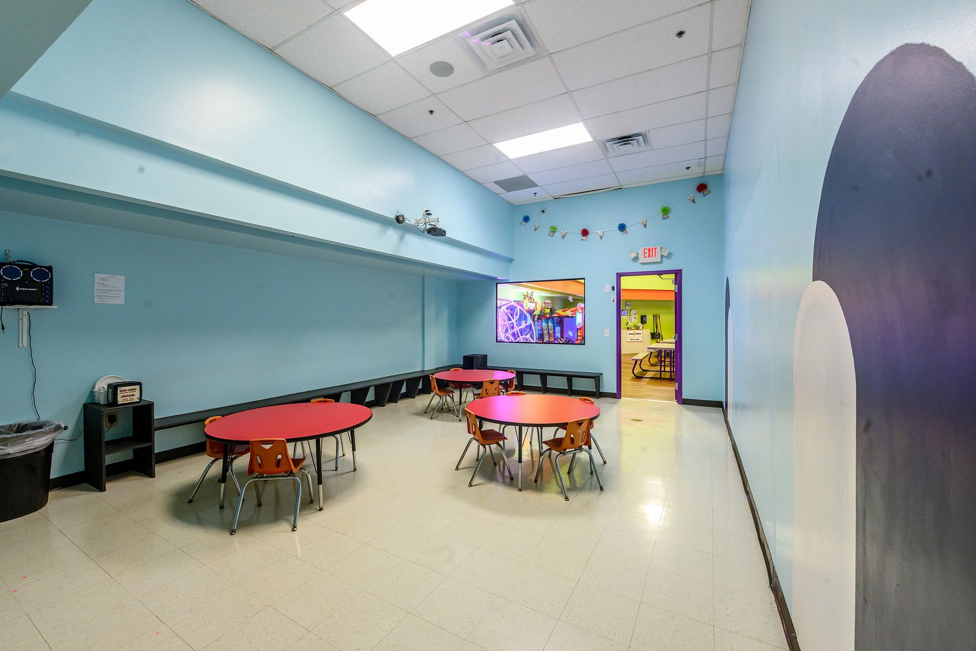 Children's play area with round red tables, small chairs, a large screen, and light blue walls.