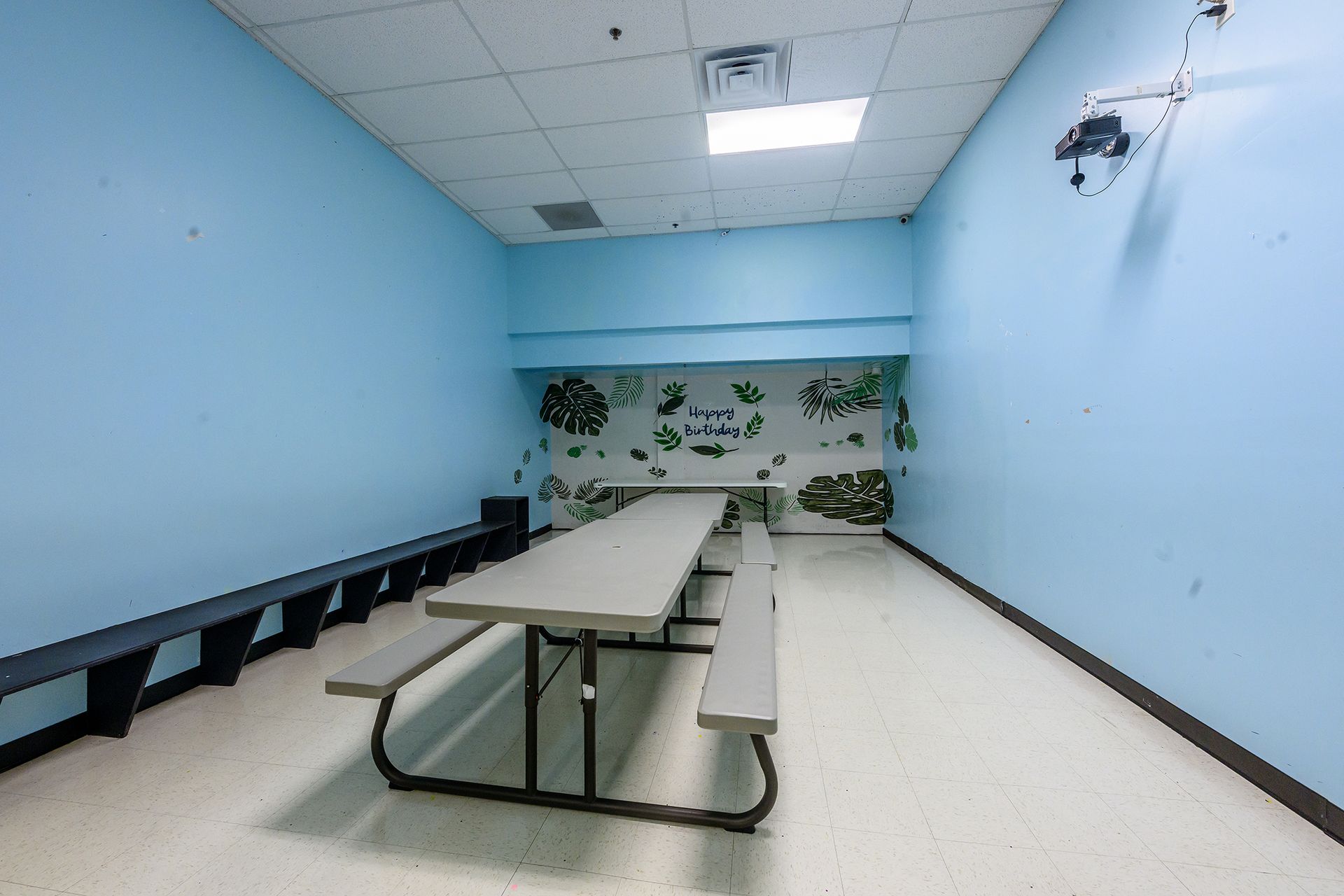 Empty room with picnic table, benches, and mural. Blue walls, white floor, fluorescent lights.