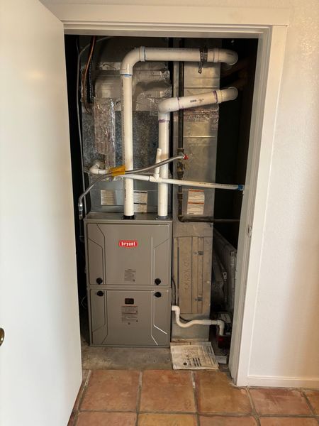 A furnace in a closet, with white pipes and ducts. Beige metal unit sits on a tile floor.