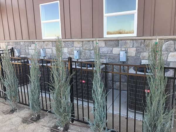 Four air conditioning units behind a black metal fence, with tall plants in front. Building with stone and windows in the background.