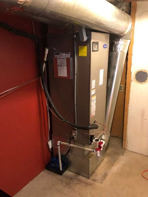A furnace and air handler unit in a basement corner, next to a red door and beige walls.