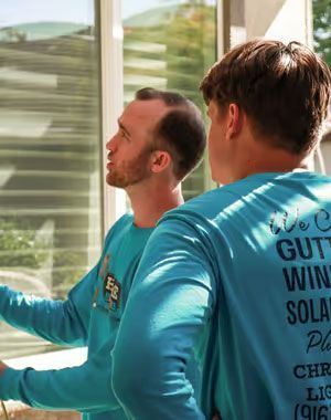 Two men in blue shirts standing by a sunlit window, one reaching toward the glass.