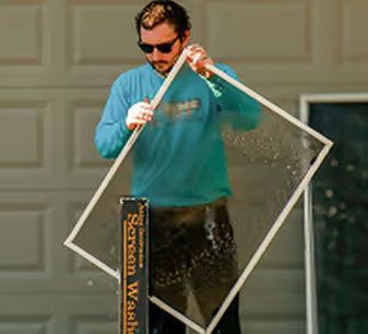 Man cleaning a window screen outdoors with a spray bottle and cloth in front of a garage door