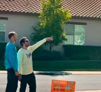 Two people stand on a street, one pointing at something off-screen near a house and orange sign.