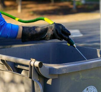 Gloved hand using a green hose to rinse a gray trash bin outdoors