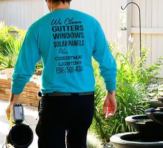 Person in teal shirt with company text stands outdoors near plants and patio decor.