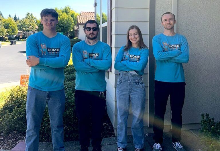 Four people in matching blue shirts stand with arms crossed outdoors beside a building.