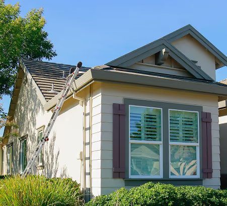 Suburban house exterior with white siding, green shutters, and a ladder on the roof under a blue sky