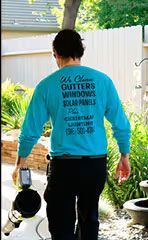 Person in teal long-sleeve shirt and black pants walking outdoors, carrying a paint sprayer and bucket.