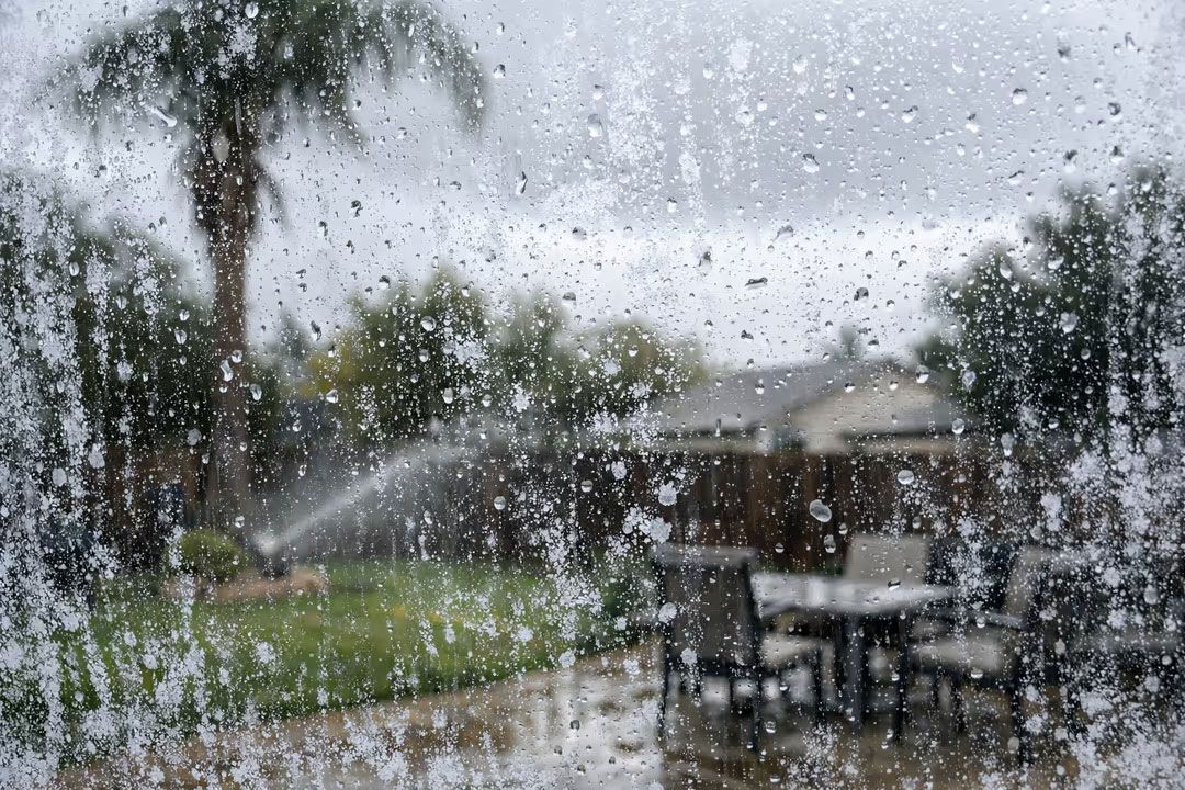 Rain-soaked patio seen through a window covered in raindrops, with tables and a spraying lawn beyond.