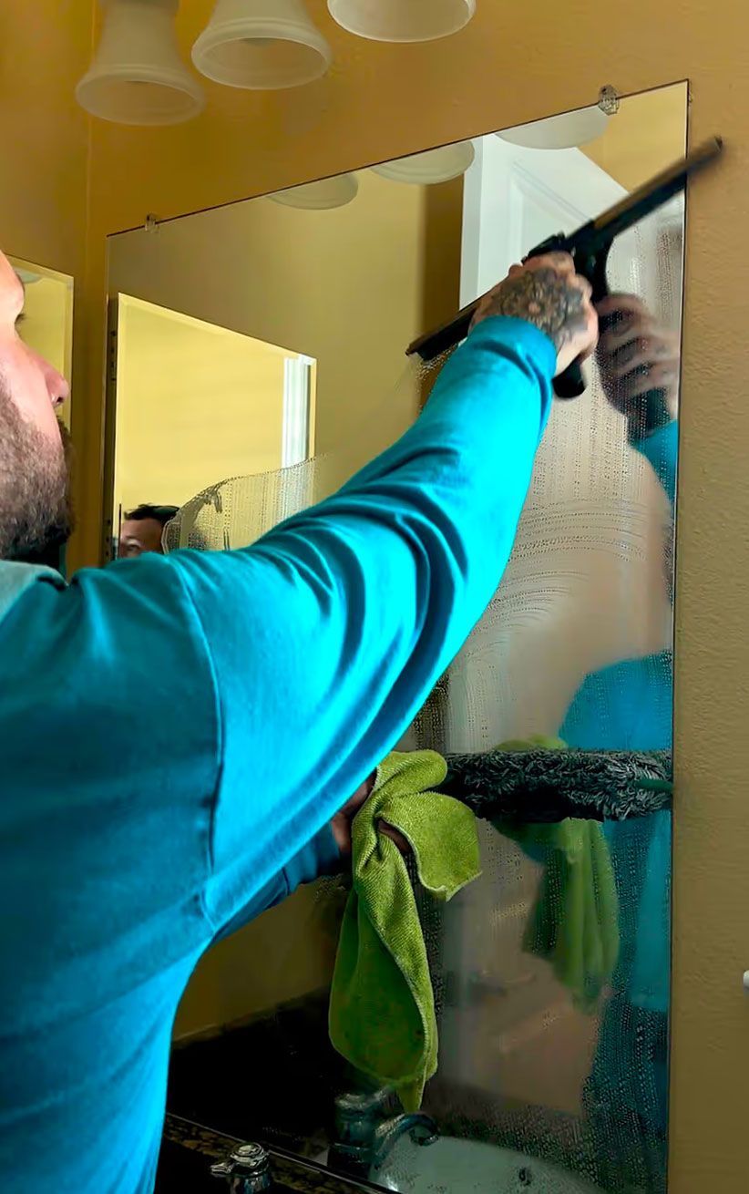 Person cleaning a bathroom mirror with a squeegee, wearing a teal shirt and black gloves.