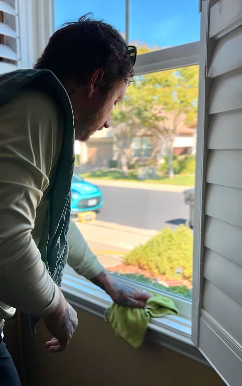 Person cleaning a white window frame with a green cloth while looking outside at a sunny street