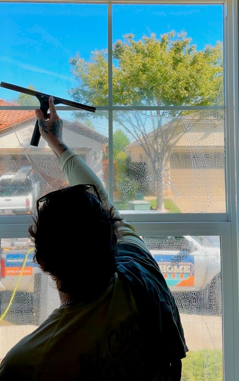 Person reaching up to clean a window, with bright blue sky and a tree visible outside.