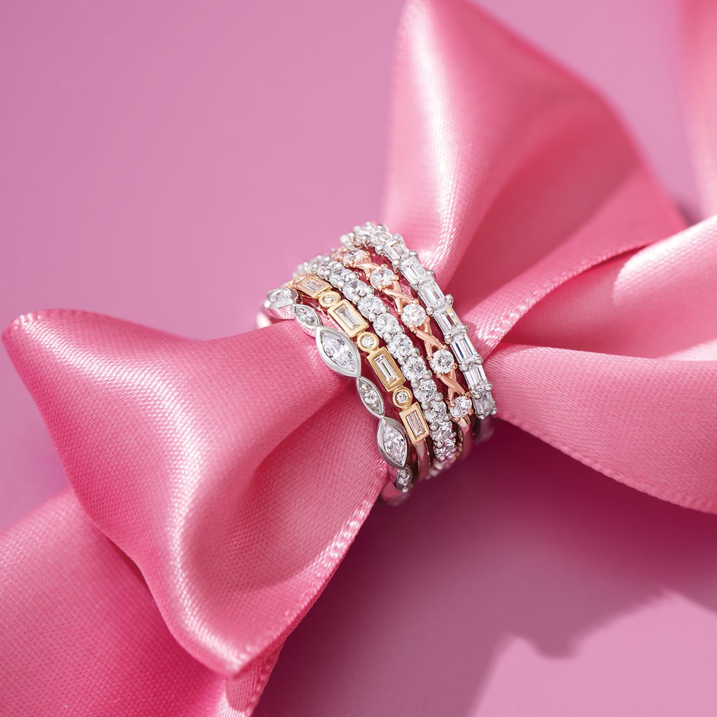 Stack of diamond rings on a pink satin ribbon against a pink background.