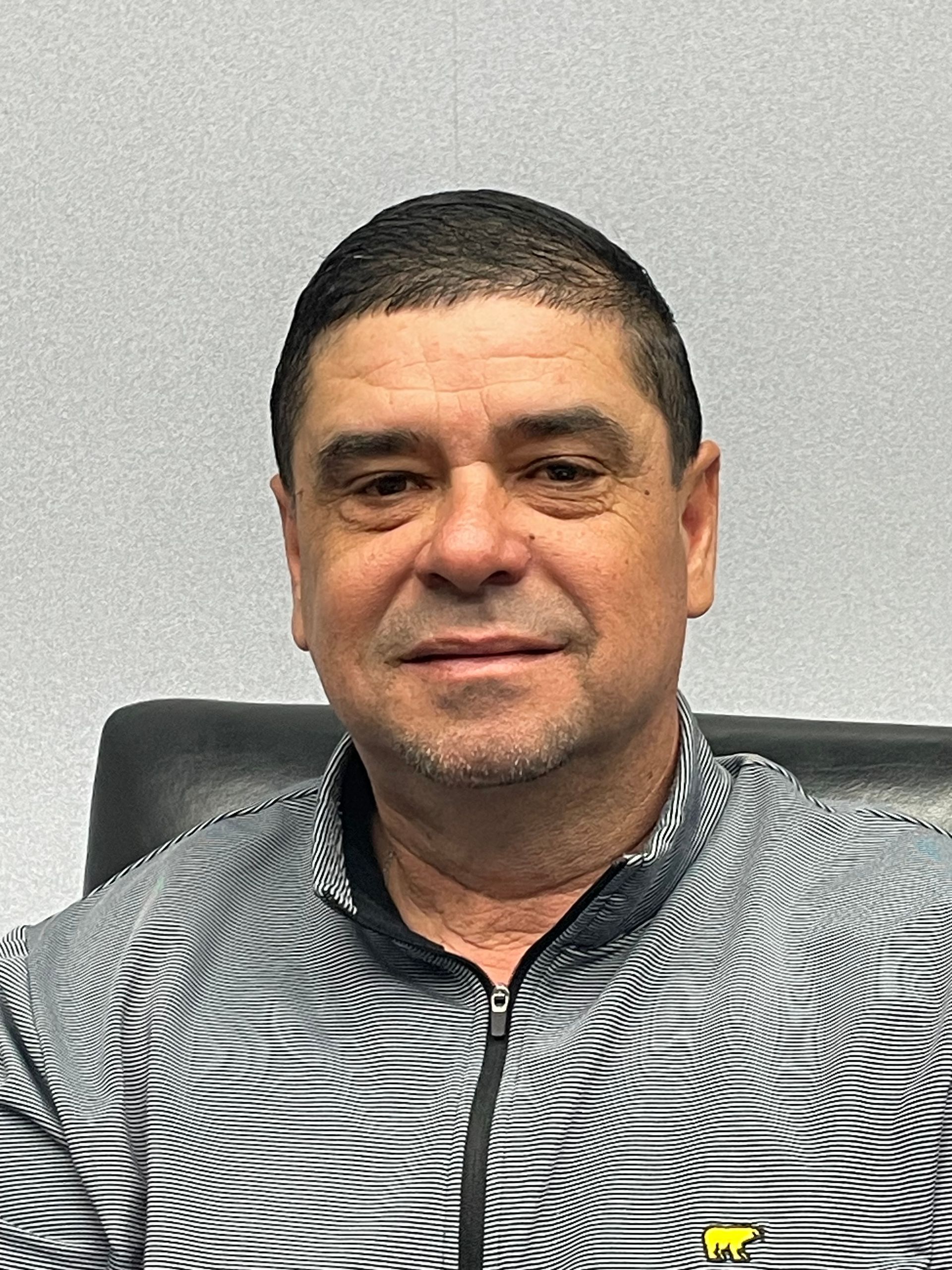 Man seated indoors against a plain wall, wearing a black-and-white patterned shirt, looking at the camera.