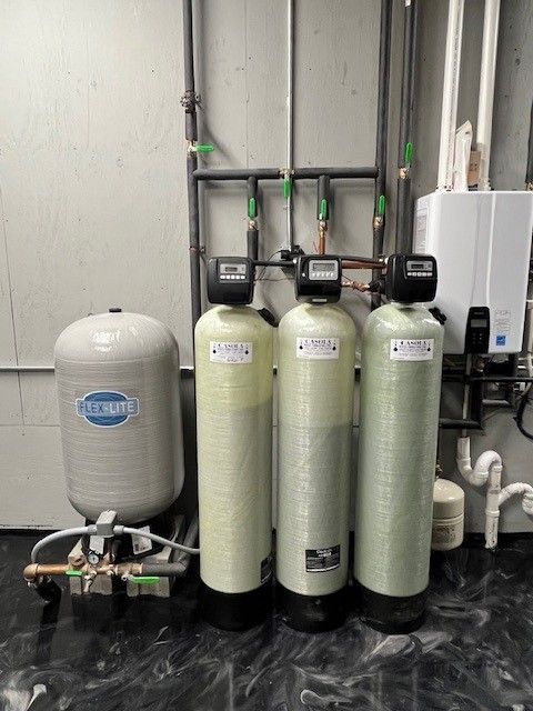 Water filtration system with three cylindrical tanks, a pressure tank, and pipes against a gray wall. 