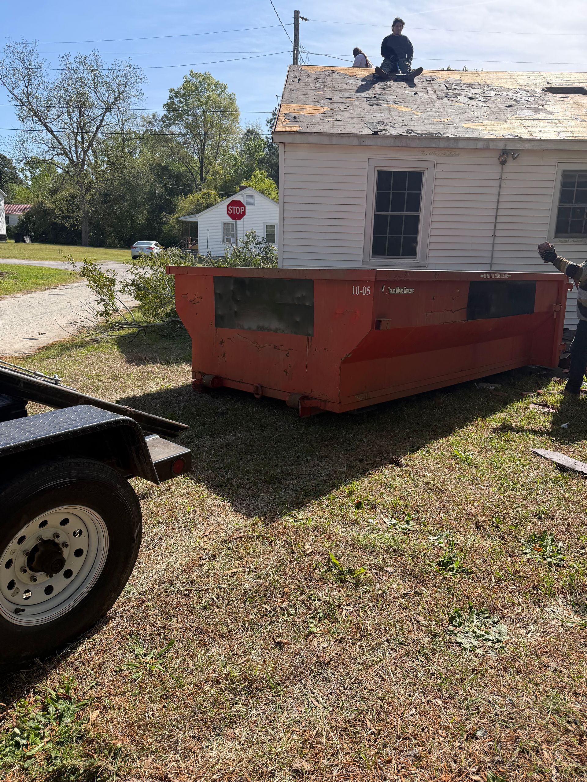 Red dumpster beside a small white house, with a worker on the roof and a truck wheel in the foreground.