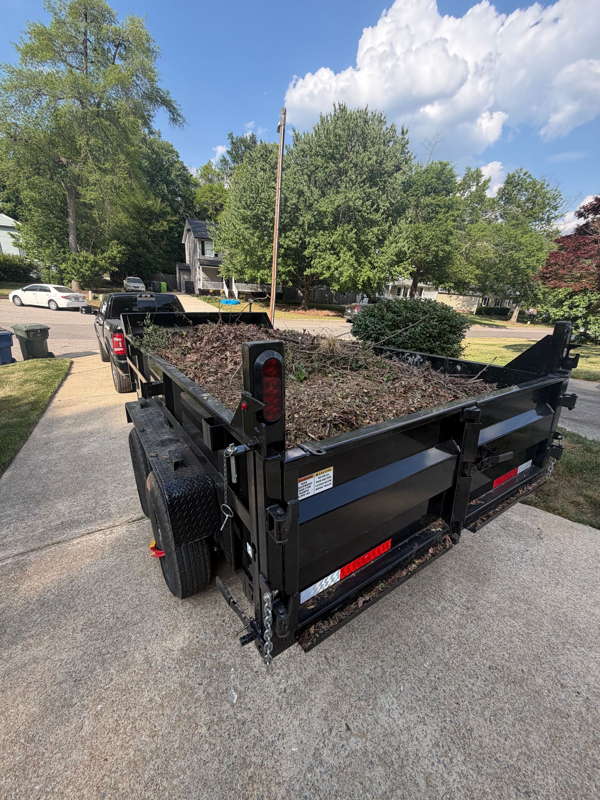 Black utility dump trailer filled with mulch parked on a gravel driveway beside trees and a house